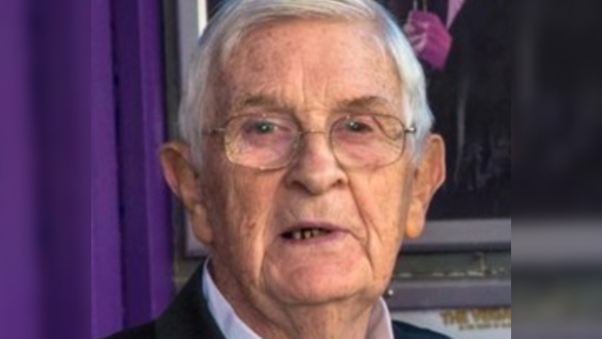 Bart O'Hare is posing for a photo in front of a purple curtain backdrop. He has short white hair and is wearing a pair of glasses, and a suit.