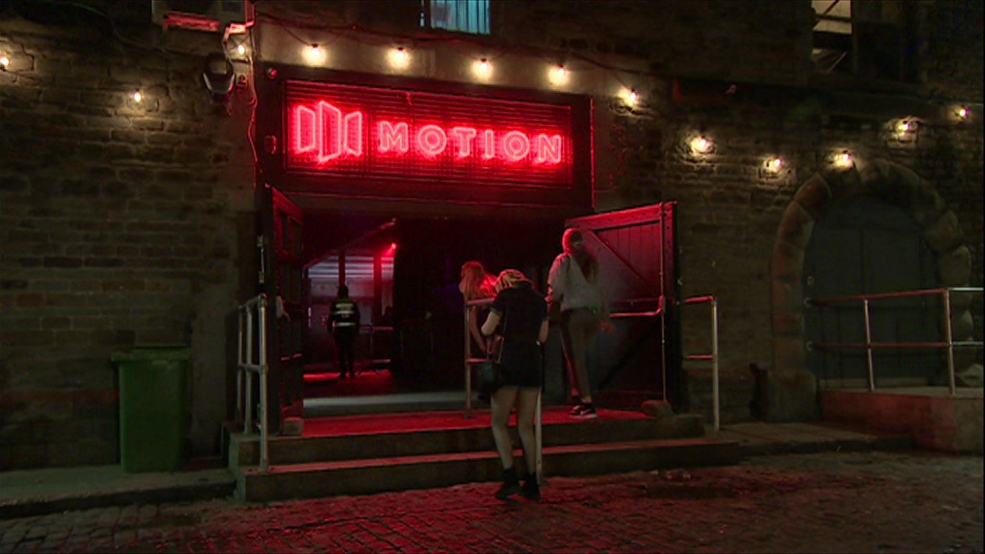 The entrance of Motion, with the word in red lights and a logo next to it. Women are walking in through the doors and it is night time.