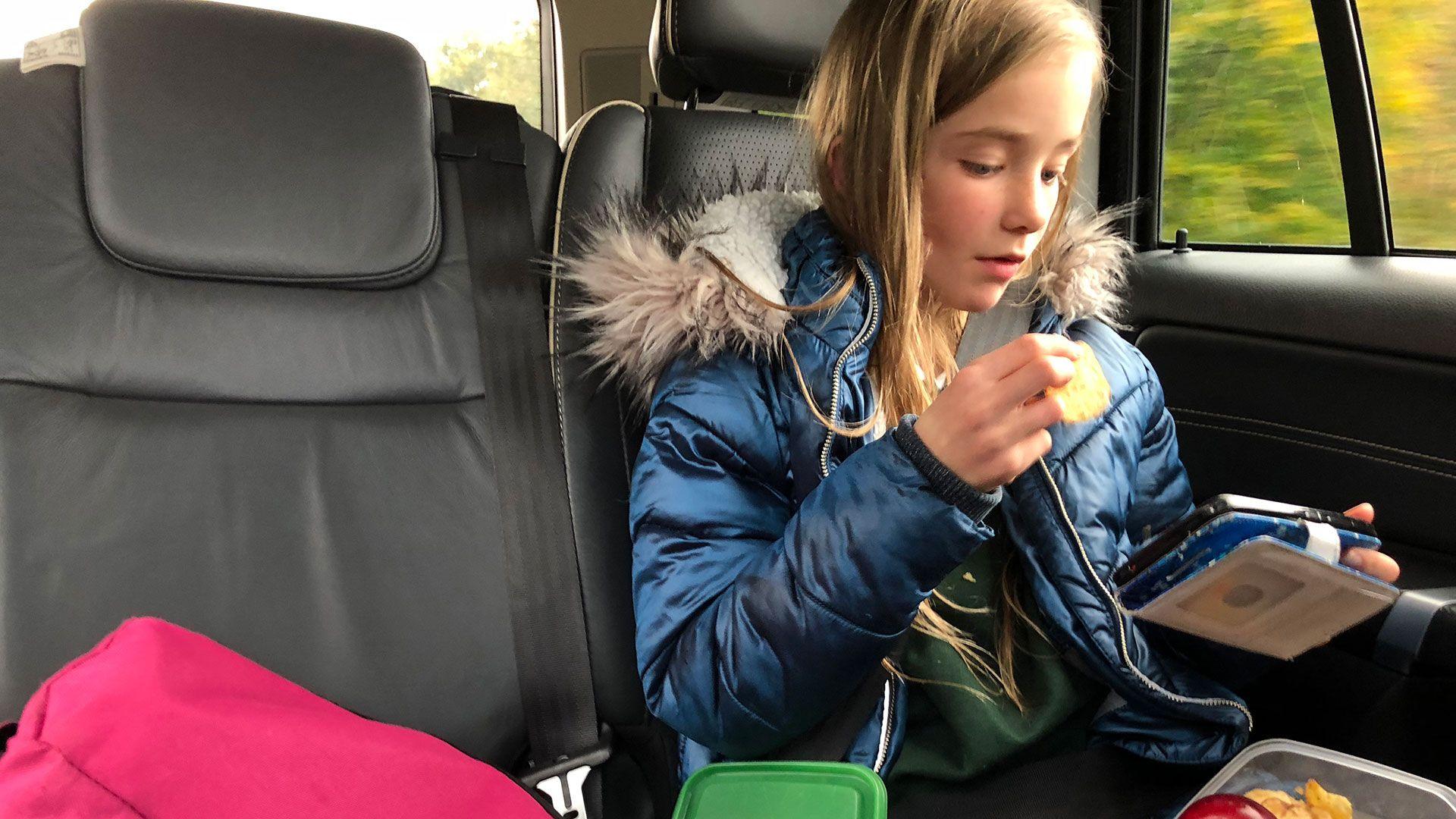 A child sits in the back on a car looking at a hand held electronic device and eating a snack from their lunch box. She is wearing a school uniform and winter coat