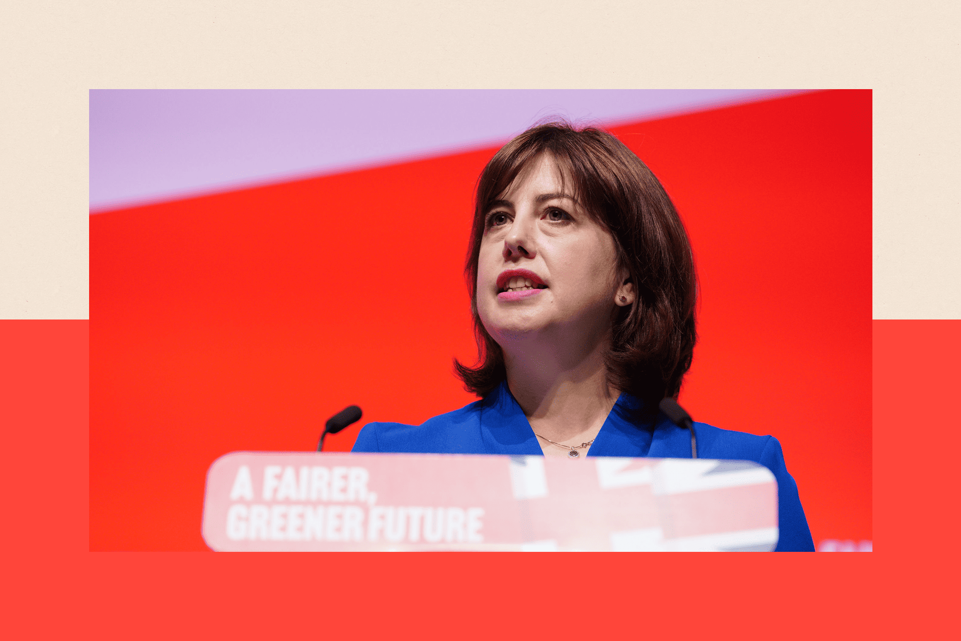 A close up of Lucy Powell, standing at a Labour-themed lectern with the union jack flag printed on it, wearing a blue blazer and in front of a bright red background.