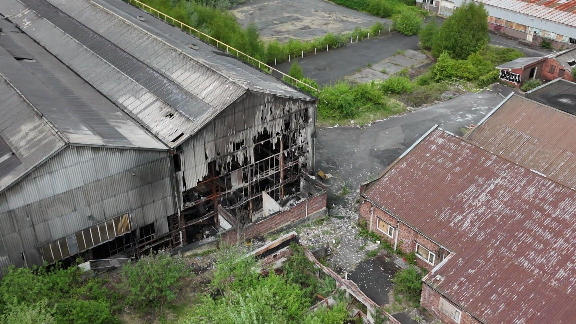 Pictures of abandoned buildings and fire damage. Two buildings that have sustained fire damage are made from corrugated iron and have corrugated iron roofs. It looks like a former factory unit. The front of the buildings have been destroyed by fire. They are in front of a boarded up brick building. The area surrounding it is overgrown with bushes and trees and looks abandoned.
