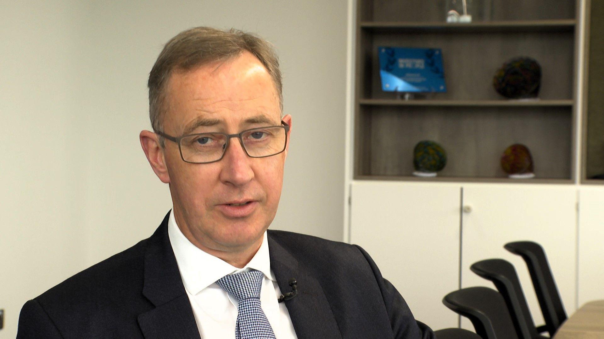 Andrew Evans, dyn busnes mewn siwt a sbectol, yn eistedd mewn swyddfa.