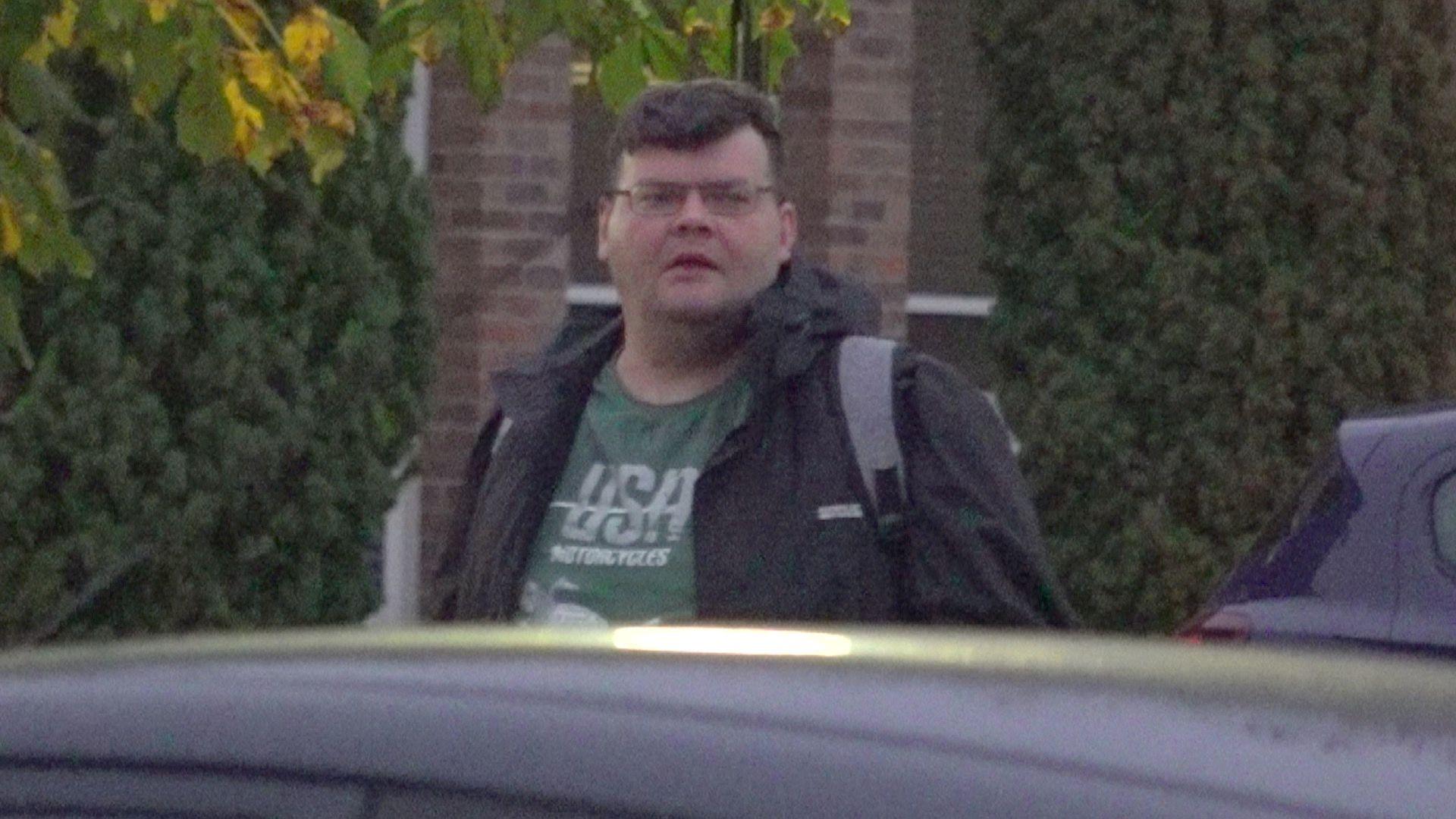 Daniel Russell looking at the photographer over the roof of a car as is outside Norwich Crown Court. He is wearing a coat and T-shirt and has a strap over his left shoulder.