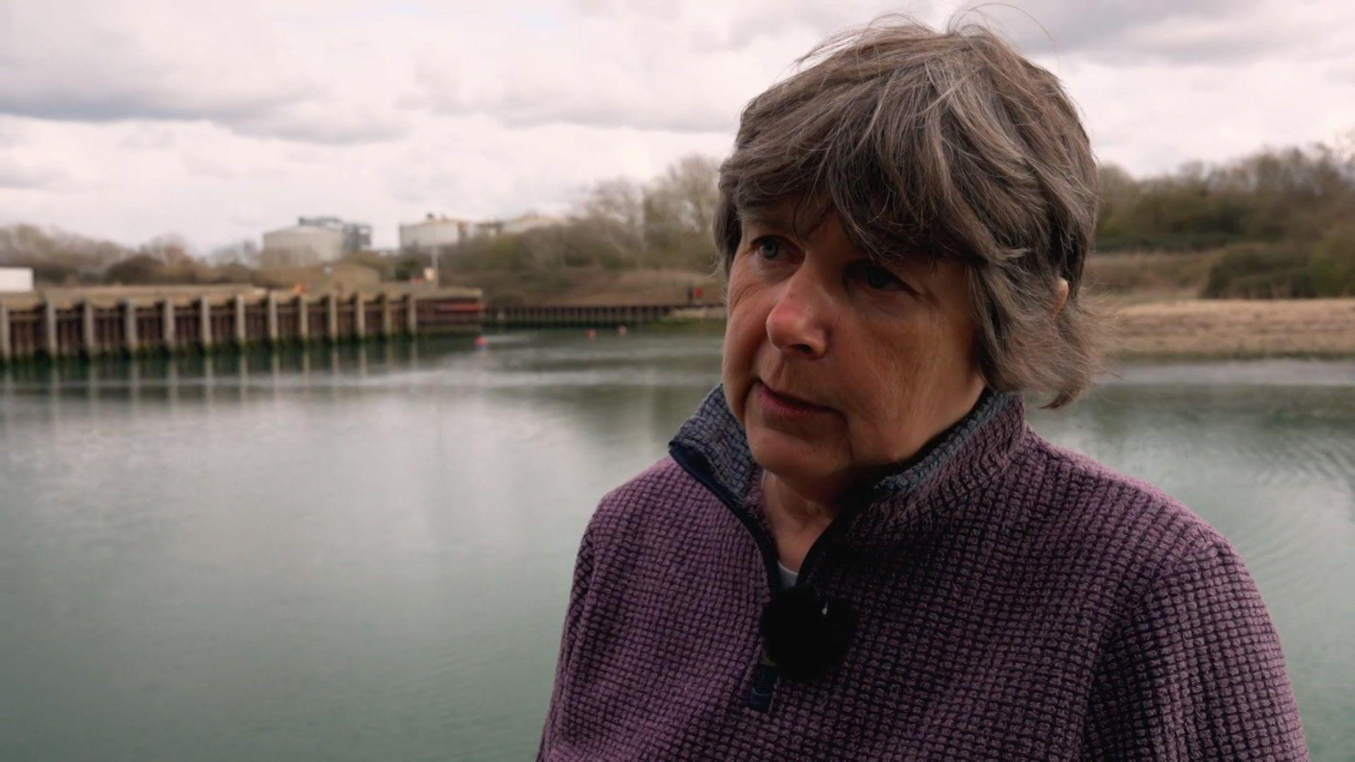 Tracey Viney stands in front of a section of water at Langstone Harbour giving an interview. She looks slightly off camera and stands to the right of the image. Tracey has short brown and grey hair, with a fringe framing her face and wears a purple textured fleece, which has a zip up collar. 