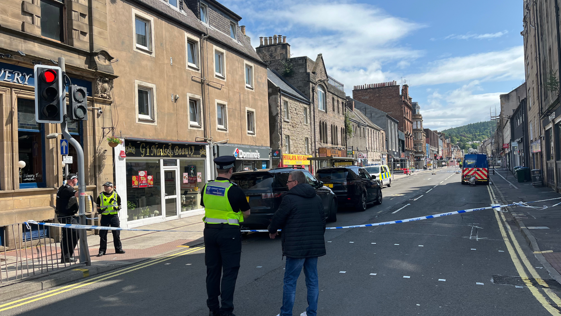 A police cordon on the street in Perth. A police officer in a hi-viz vest is speaking to a man in a dark jacket and jeans. They are standing in front of a strip of police tape.