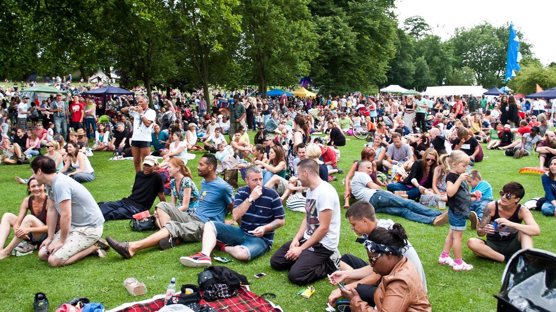 A crowd of people, some sitting, others standing, in a lush green grass in a bright day, during a summer festival
