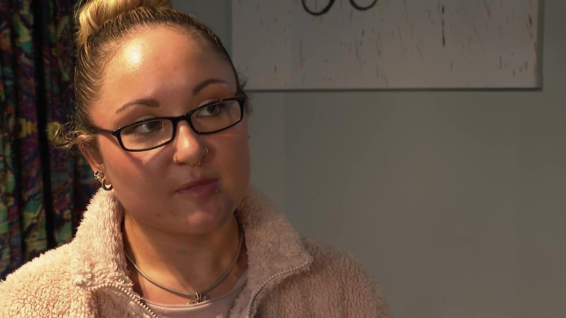 A young woman, wearing black rimmed glasses and a white fluffy jacket, has earrings, a necklace and nose piercings. She wears her dark blonde hair in a bun.