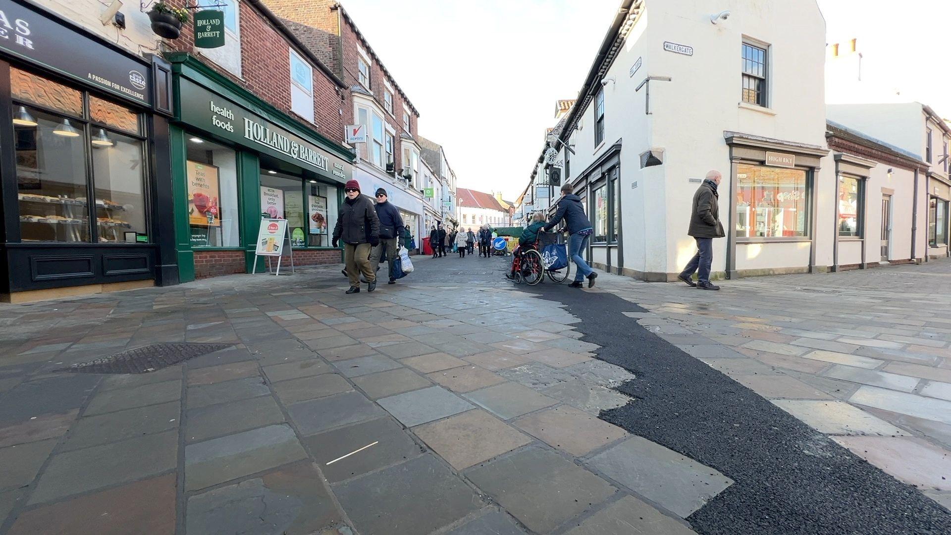 A patio pedestrianised shopping street with shops on either side and people walking.