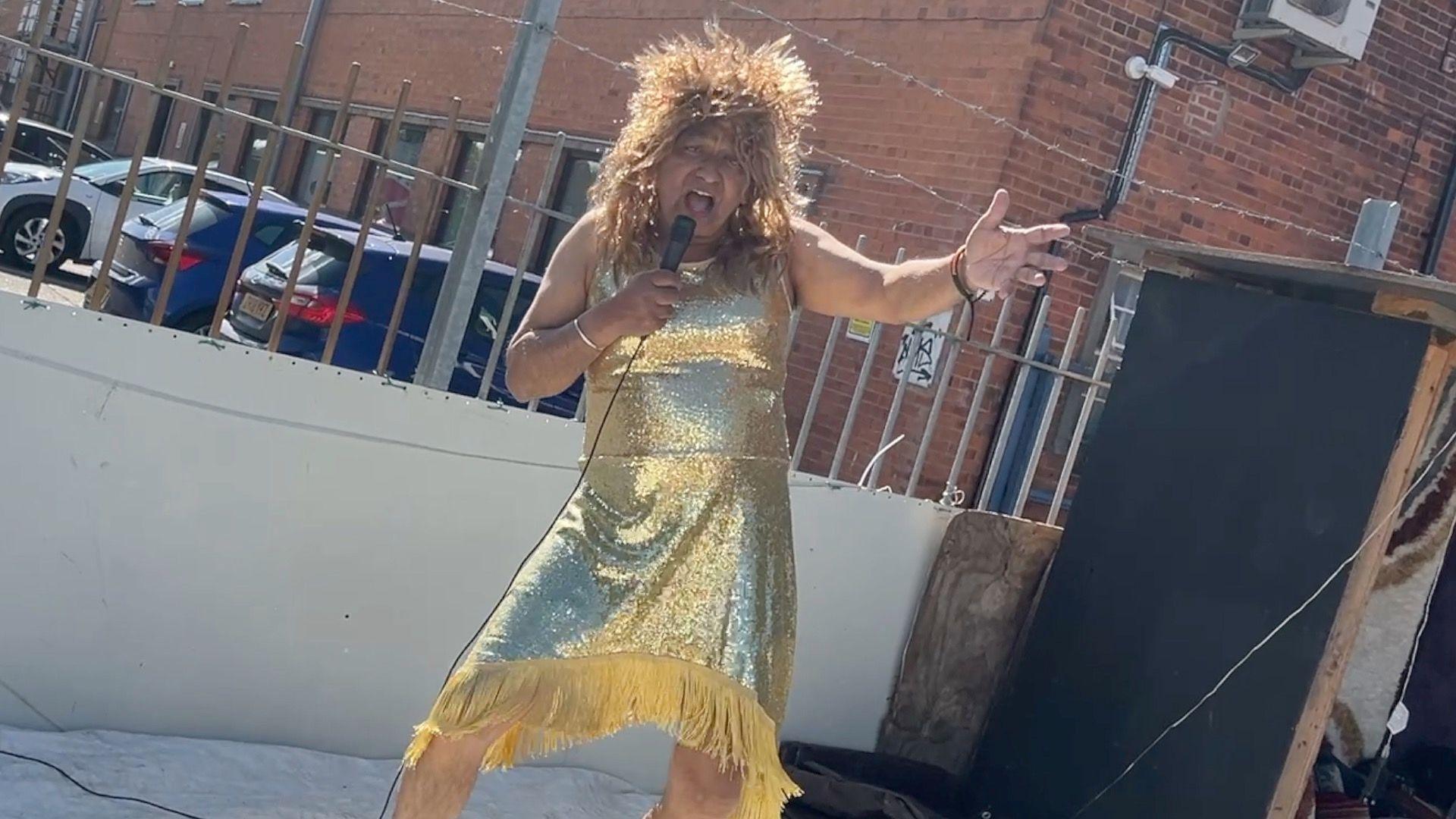 Man in a gold glittery dress with a mircophone singing on a stage outside his shop. 