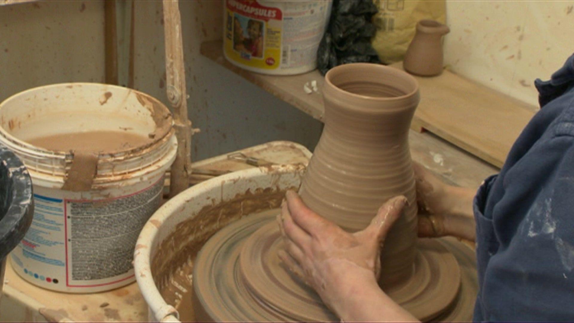 A man wearing a blue top touching some wet clay and shaping it with his hands whilst spinning it round