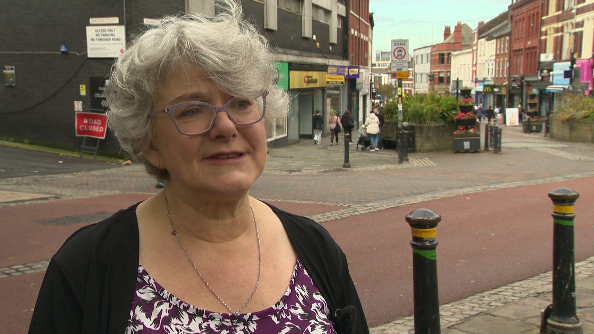 Woman with short grey hair and glasses standing in a street in Preston.