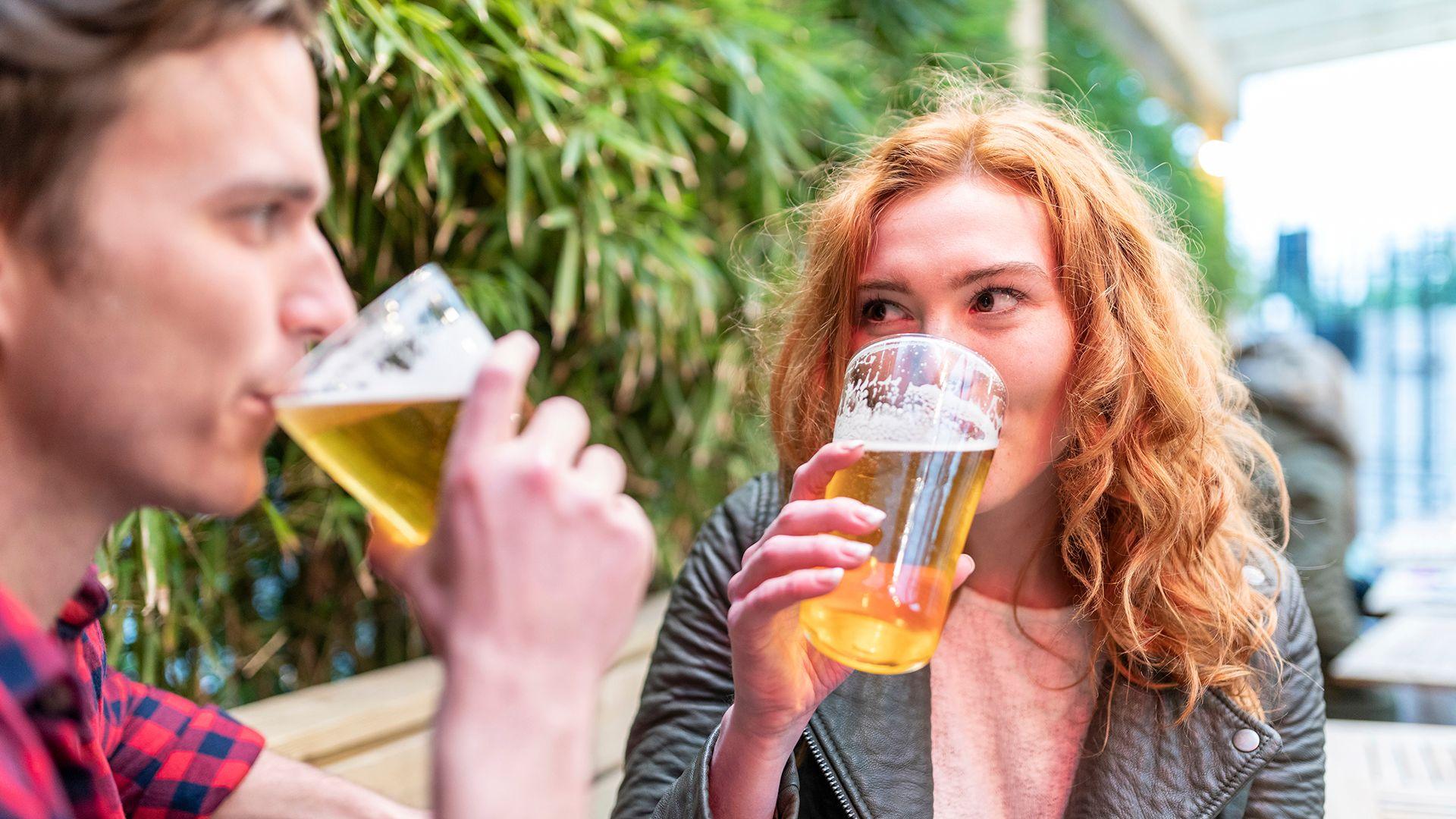 Stock photo shows two people sitting outside drinking pints of beer in front of pub garden foliage, one has short brown hair, the other light ginger hair.