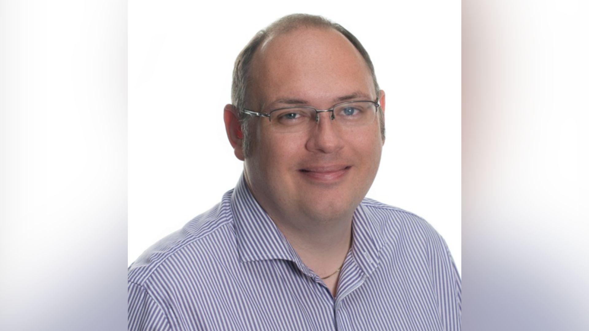 A head and shoulders picture of a white man with thinning hair.  He is smiling a the camera and wearing a blue and white thinly stripped shirt and glasses