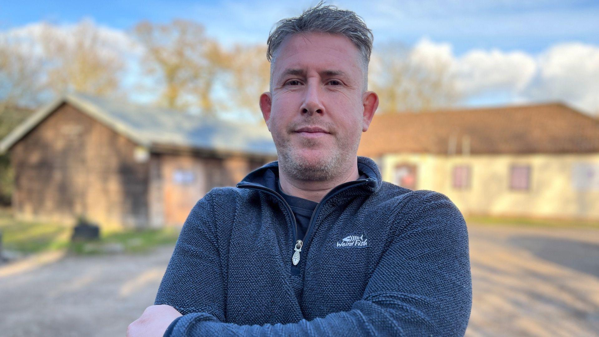 Dan Land, the chairman of Thorpe-le-Soken Parish Council, standing in front of the Scout hut in the village which has been closed. He is wearing a navy blue quarter-zip fleece top with the Weird Fish logo on his left chest. He is stood with his arms folded.