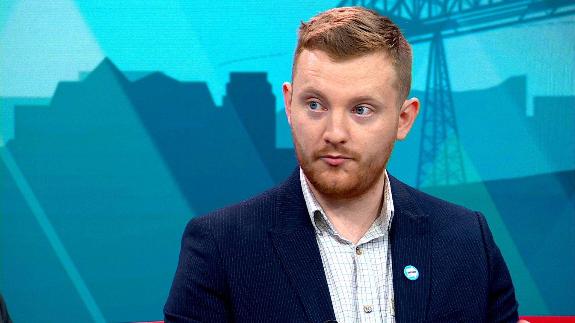 Chris Eynon sits with the teal backdrop of the BBC Politics North set behind him. He's wearing a navy pinstripe suit and checked shirt. He has short ginger hair and beard.