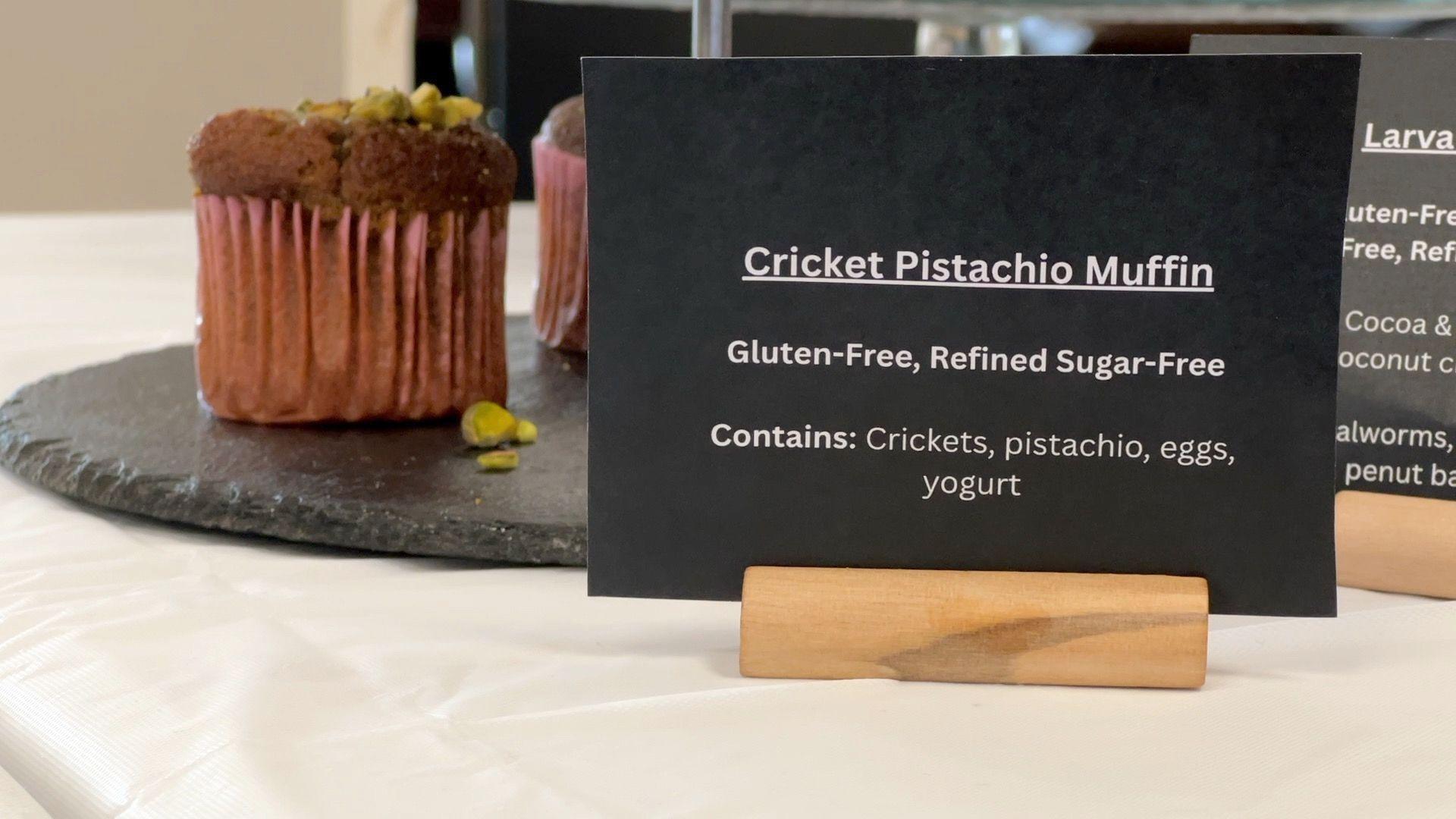 A small black sign detailing the ingredients of a Cricket Pistachio Muffin. The sign reads "Cricket Pistachio Muffin. Gluten-free, refined sugar-free. Contains: Crickets, pistachio, eggs, yoghurt"