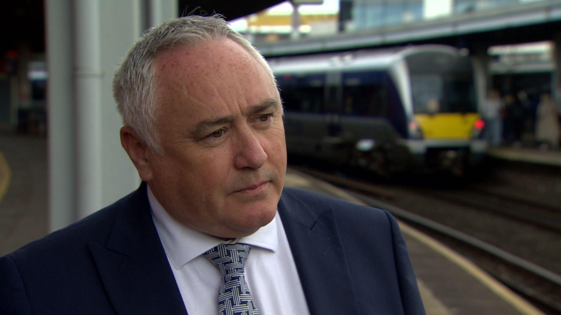 David Curry stands on a train platform, with train in the background, wearing a navy suit, white shirt and patterned  navy tie