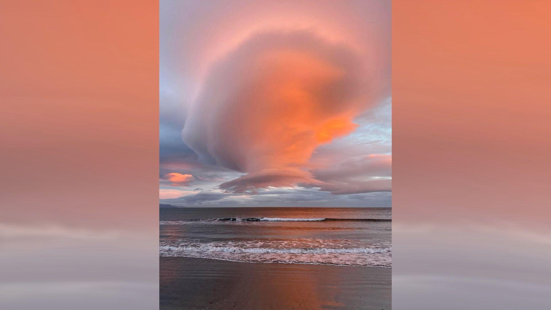 A cone shaped cloud seen in the sky over Ballygally beach. The image shows the waters edge, and a small wave coming in towards the beach. 