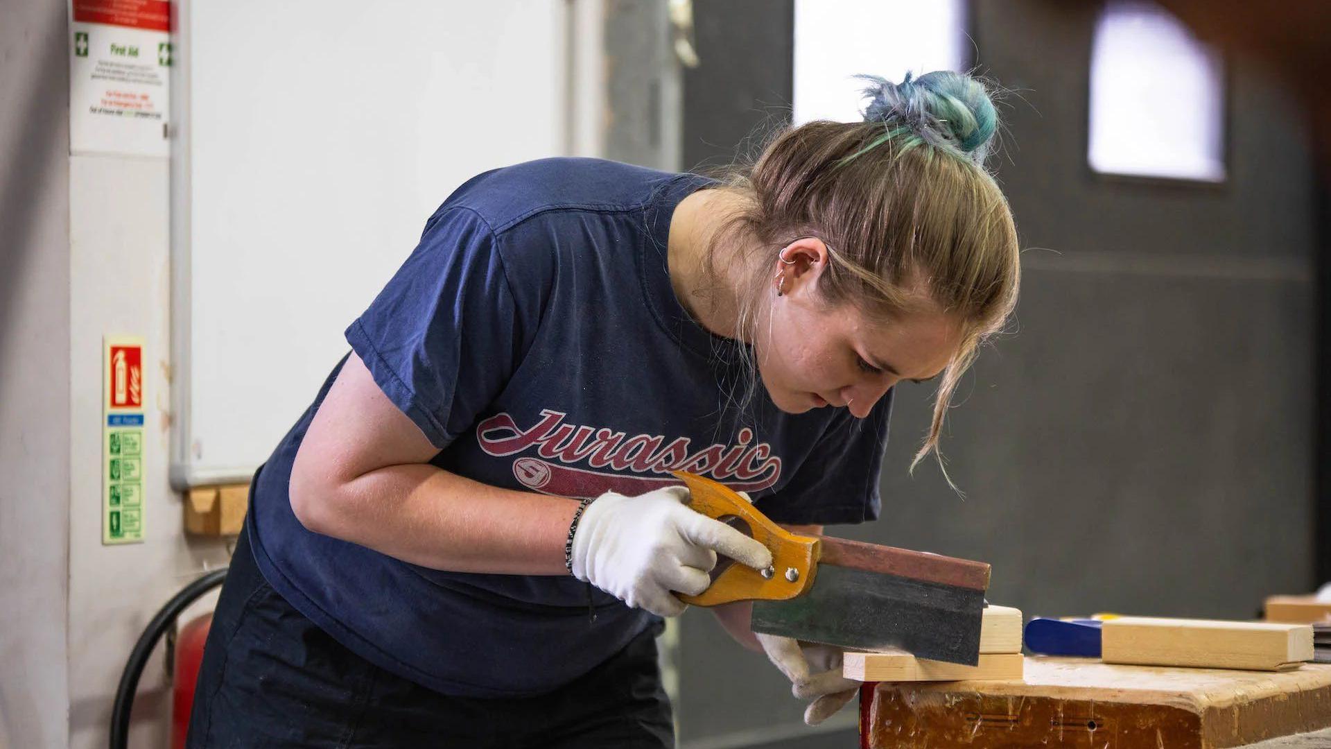 A young woman sawing a piece of wood. She is wearing a navy blue T-shirt and white gloves and is looking at the wood intently.