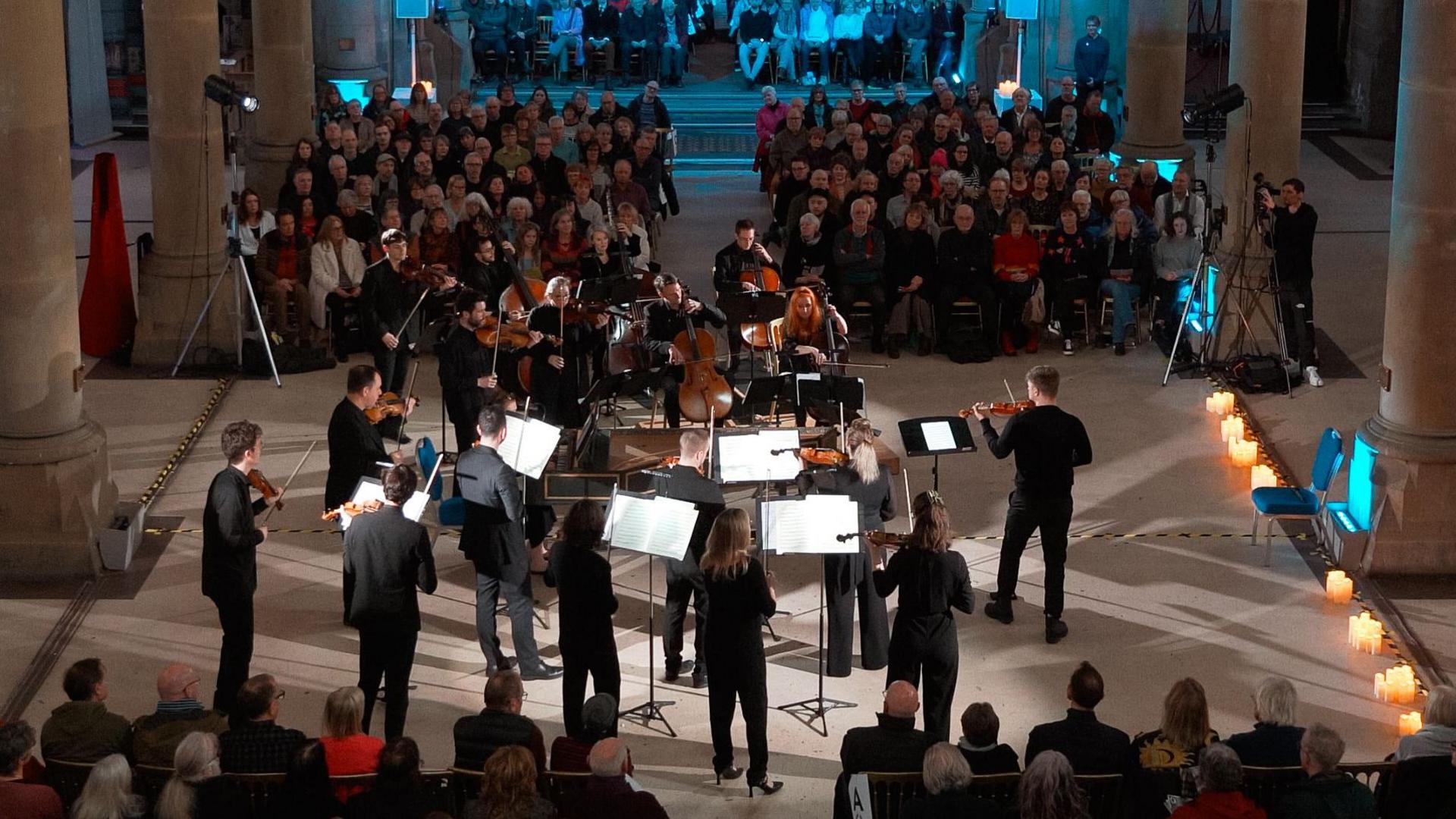 An orchestra plays string instruments. Sitting on either side of them are audience members sitting in wooden chairs. They are all gathered inside a monastery.