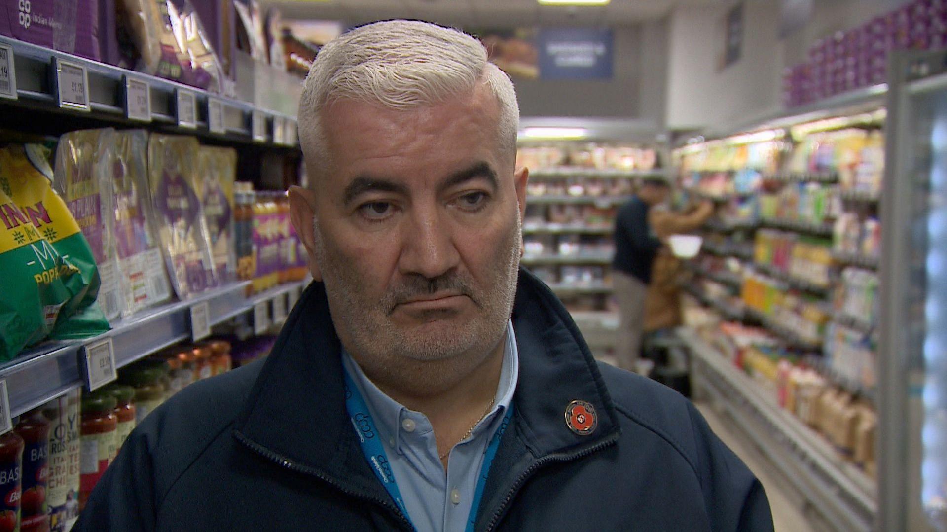 Mark Cox looks serious. He is standing in a supermarket aisle with a chiller on one side and packed shelves on the other. He is wearing a blue coat over a blue shirt and a remembrance poppy.