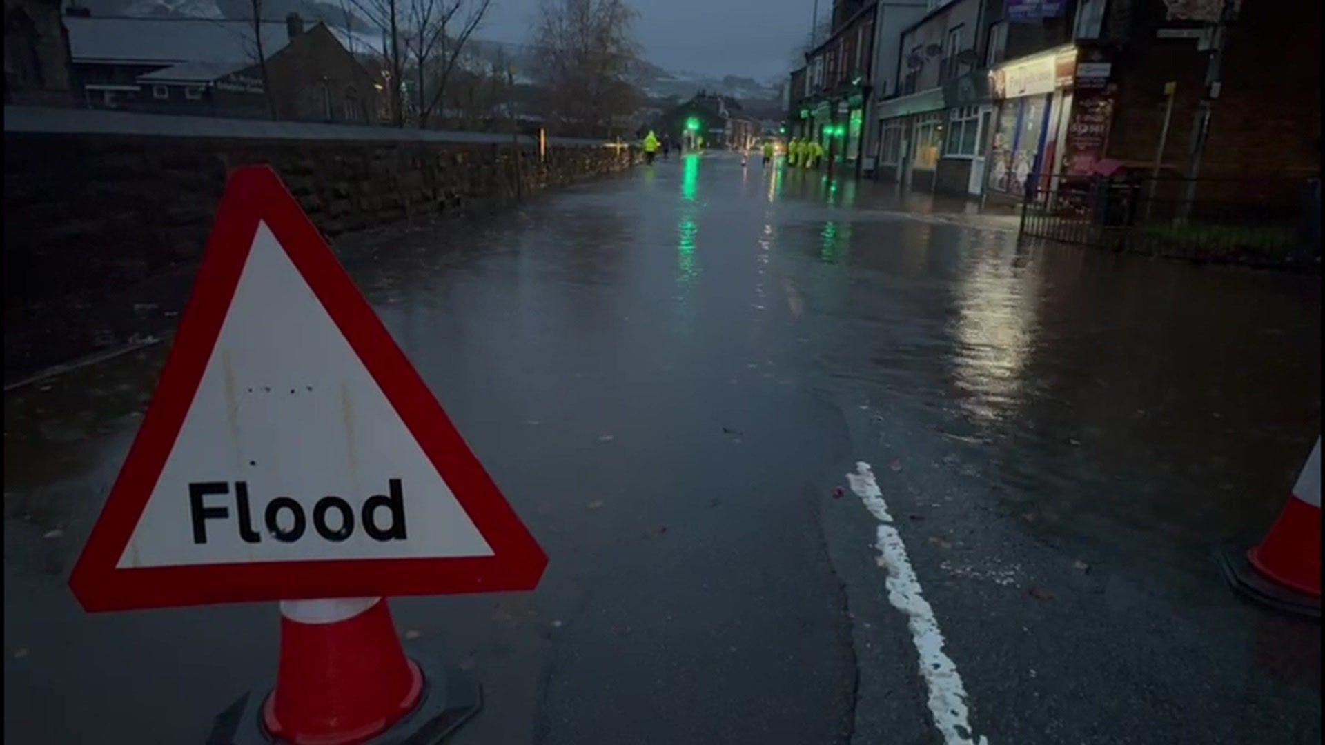 A red and white triangle sign that reads 'Flood' in front of a flooded street with green lights and people in yellow high-vis jackets in the distance.