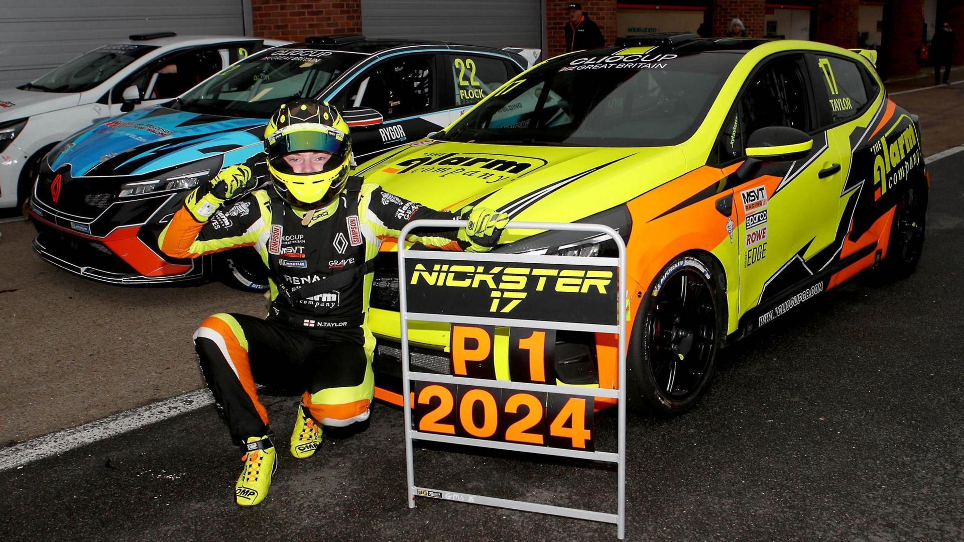 Nicky Taylor kneels to the left of a sign saying "P1 2024" to celebrate the Clio Cup GB Championship win. He's in front of his title-winning car, beside two other competing cars.