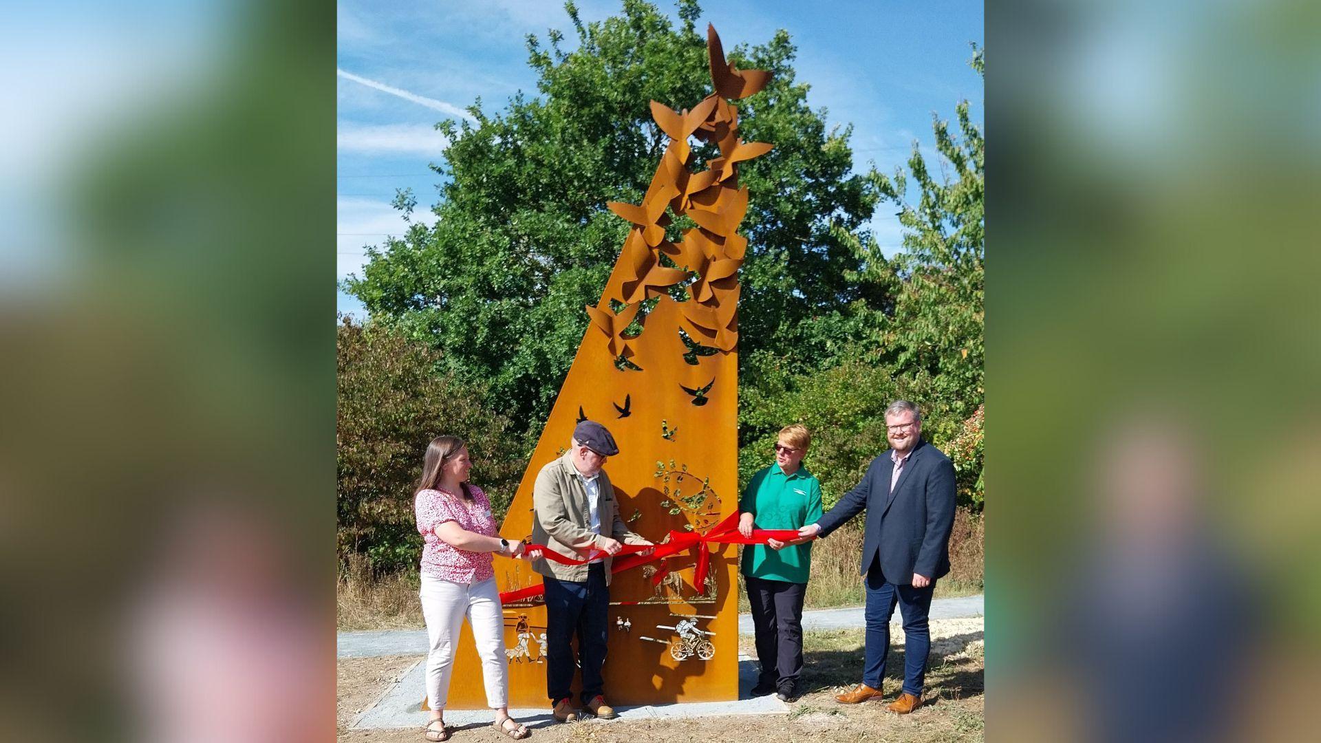 A ribbon being cut at the sculpture's unveiling. It is a tall, steel structure which has been painted bronze. At the bottom, silhouettes of families walking and cycling have been cut out. At the top, it becomes a weave of steel birds flying upwards.