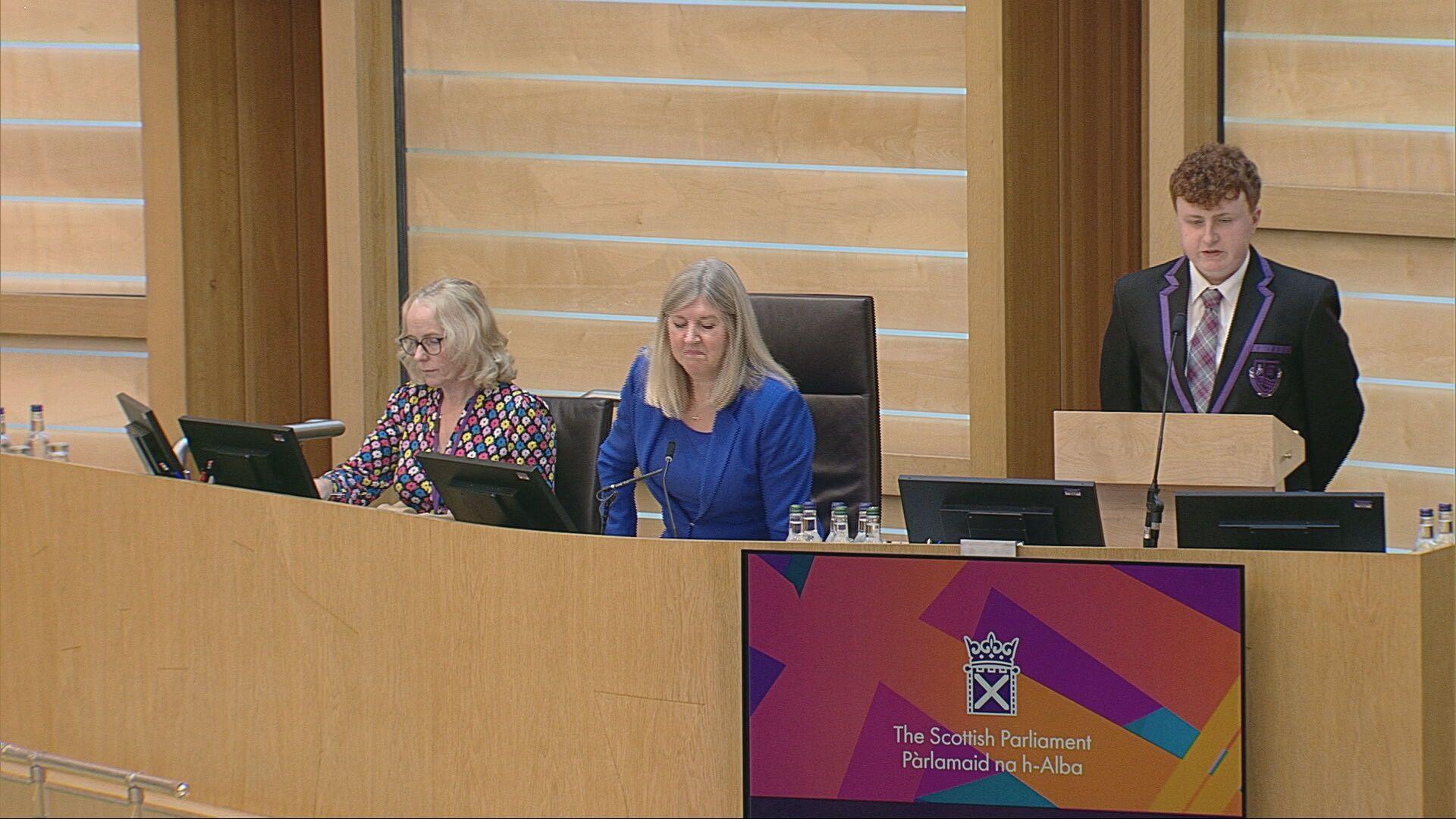 A teenage boy standing up behind a podium in a parliamentary building, wearing a black school uniform with purple trim and tartan tie. Two women are sitting along from him, one wearing a blue suit and the other in a multi-coloured top. The walls and decor of the room are beige.