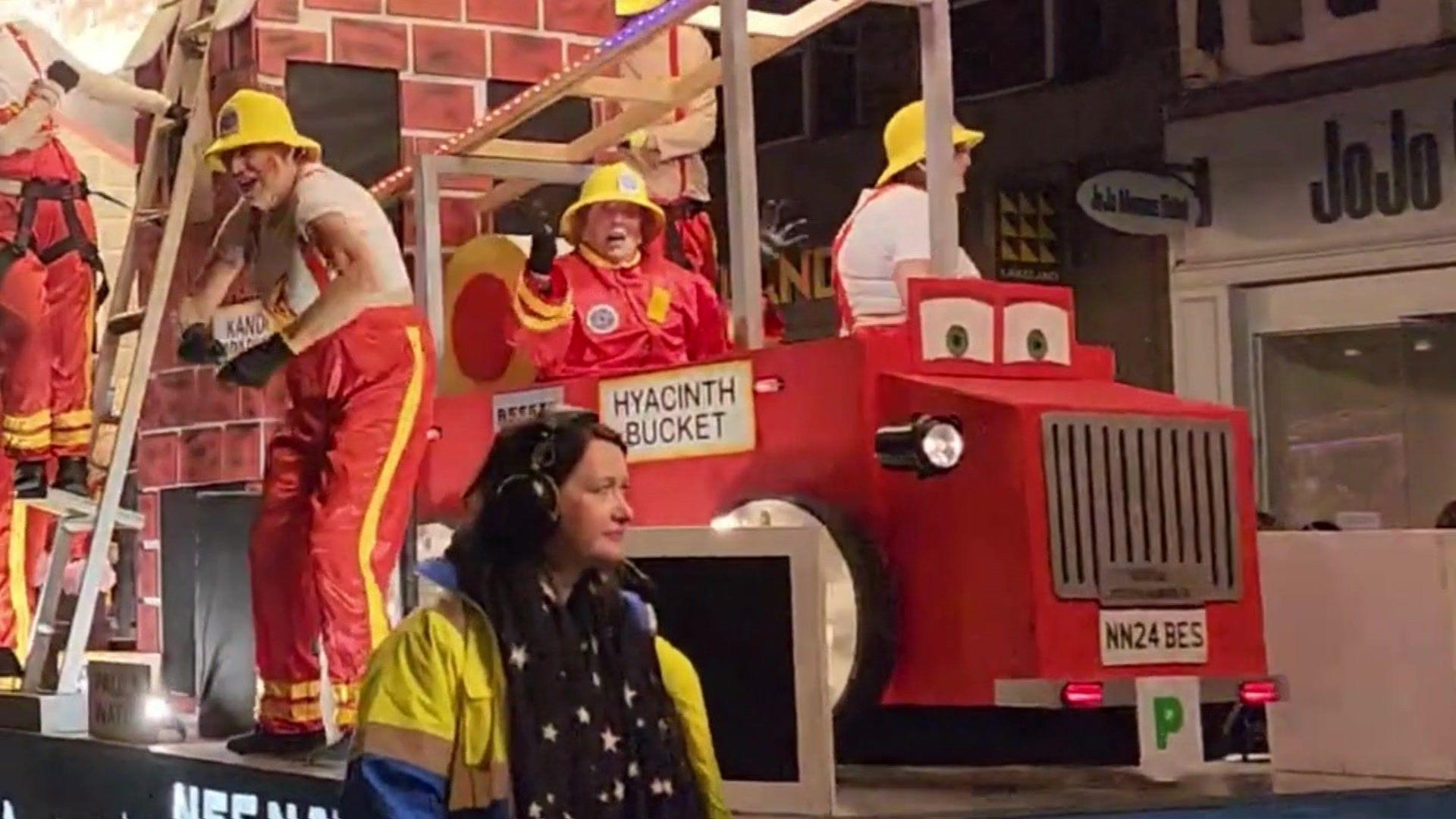 Irene Hill sits on a carnival float. She is wearing a red firefighter outfit and is sat in a prop fire engine. There are people stood on either side of her dancing for the crowd, also in firefighter outfits. In the foreground of the image a steward in a high-vis jacket wearing headphones looks off camera.