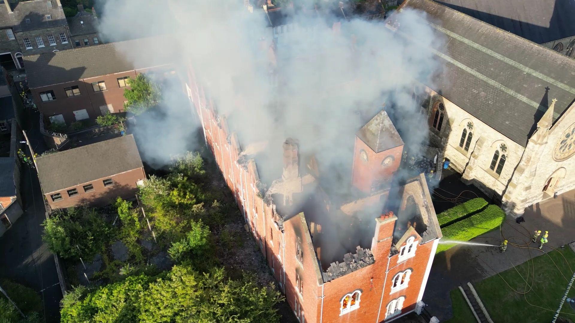 Drone footage from above of a red-brick building with flames inside it. The building is large and long with its roof caved in. It is surrounded by small red-brick buildings and a grey stone church beyond it. Firefighters in hi-viz protective clothing are spraying it with water. 
