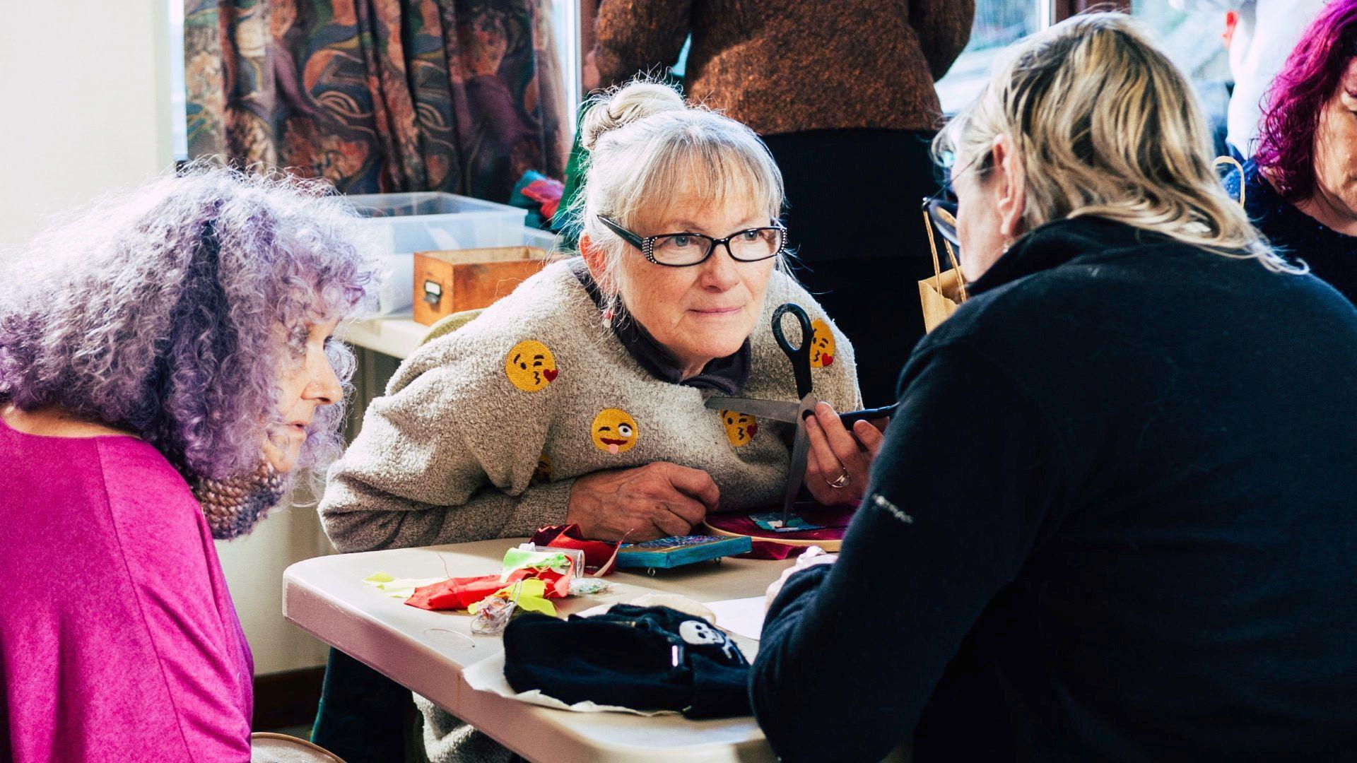 Three women are huddled around a table chatting. There are sewing materials on the table. One woman has her back to the camera, she is wearing a black fleece and has blonde hair. To her left a woman is looking at her while she speaks. This woman has purple curly hair and wears a bright pink light wool jumper. In the centre of the frame, a woman is crouched slightly over the table like she is listening intently. She has blonde/grey hair styled with a fringe and tied back into a bun. She wears rectangular framed black glassed and a beige fluffy woolen jumper, which has yellow face emojis prints scattered all over it. She is holding a pair of scissors in her hand.