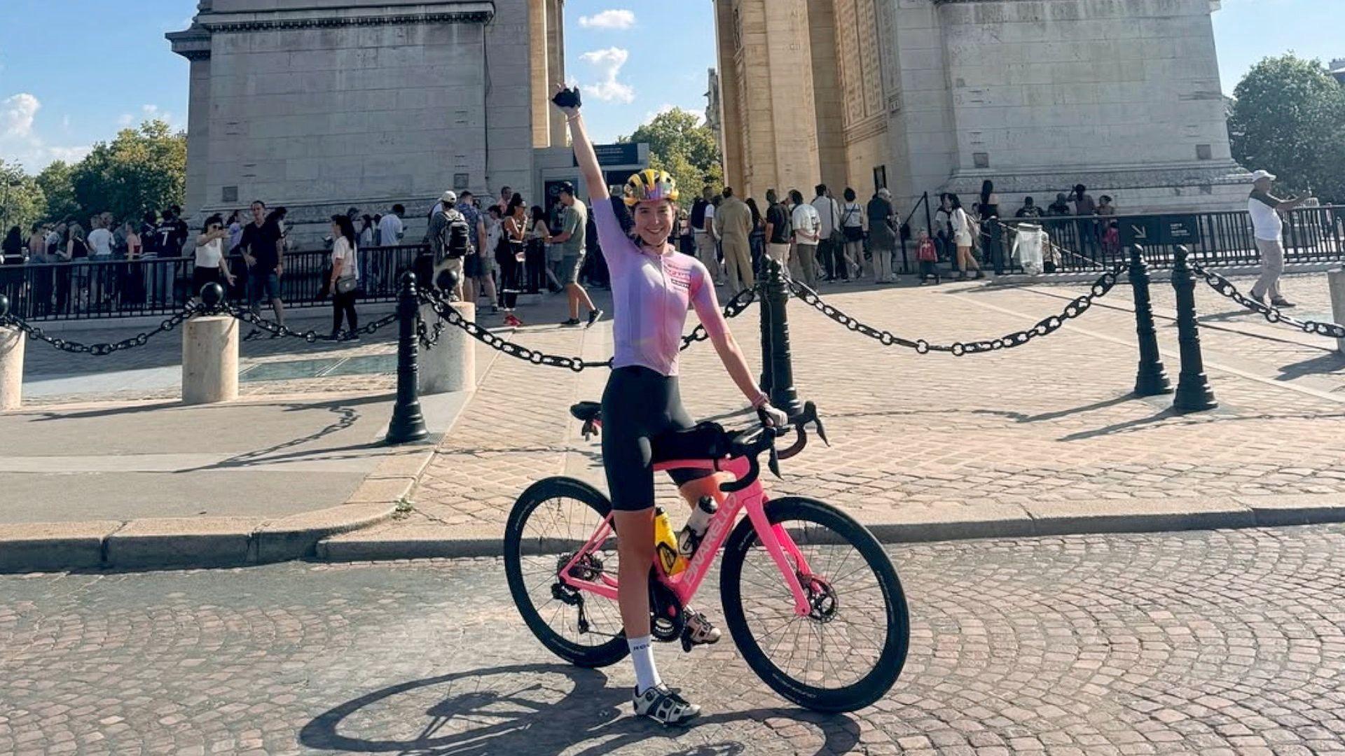 Amy Hudson at the Arc de Triomphe in Paris. She is dressed in a pink and white cycling top, black shorts and a yellow helmet. She is sitting on her pink bike with her arm pointing in the air, with the base of the Arc de Triomphe behind her in light coloured stone.
