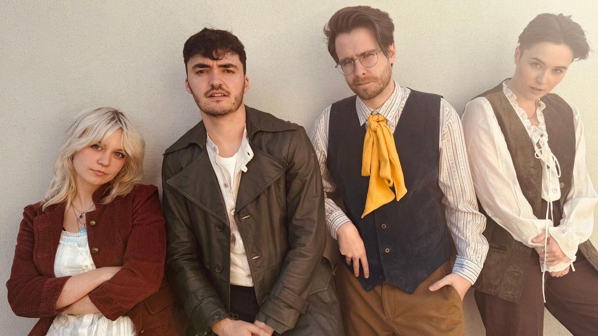 Four people line up against a wall in old fashioned costumes as part of their roles in The Highwayman. From left, a blonde-haired woman has a red jacket on over a white top and has her arms folded; a man with short dark hair and stubble beard has a brown leather coat on over a white top; a man with brown hair and glasses is wearing a blue waistcoat with a yellow tie and has his left hand in his pocket; a woman with short dark hair has a brown waistcoat over a white top and is holding her hands together.