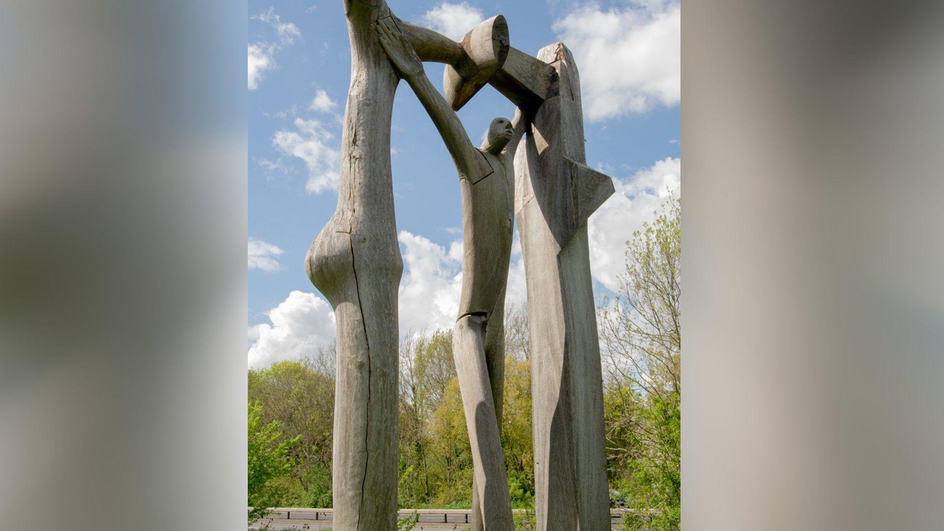 A wooden sculpture looking like a man with his hands in the air, above his head holding a wooden frame.