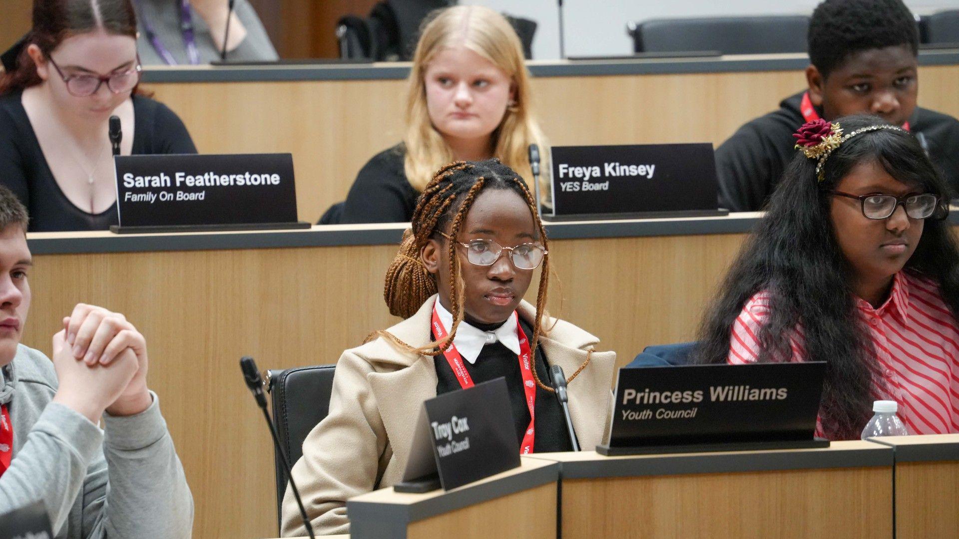 Young people sat in the council chamber. They have signs with their names on and microphones to speak in to. There are six young people in this picture, four girls and two boys.