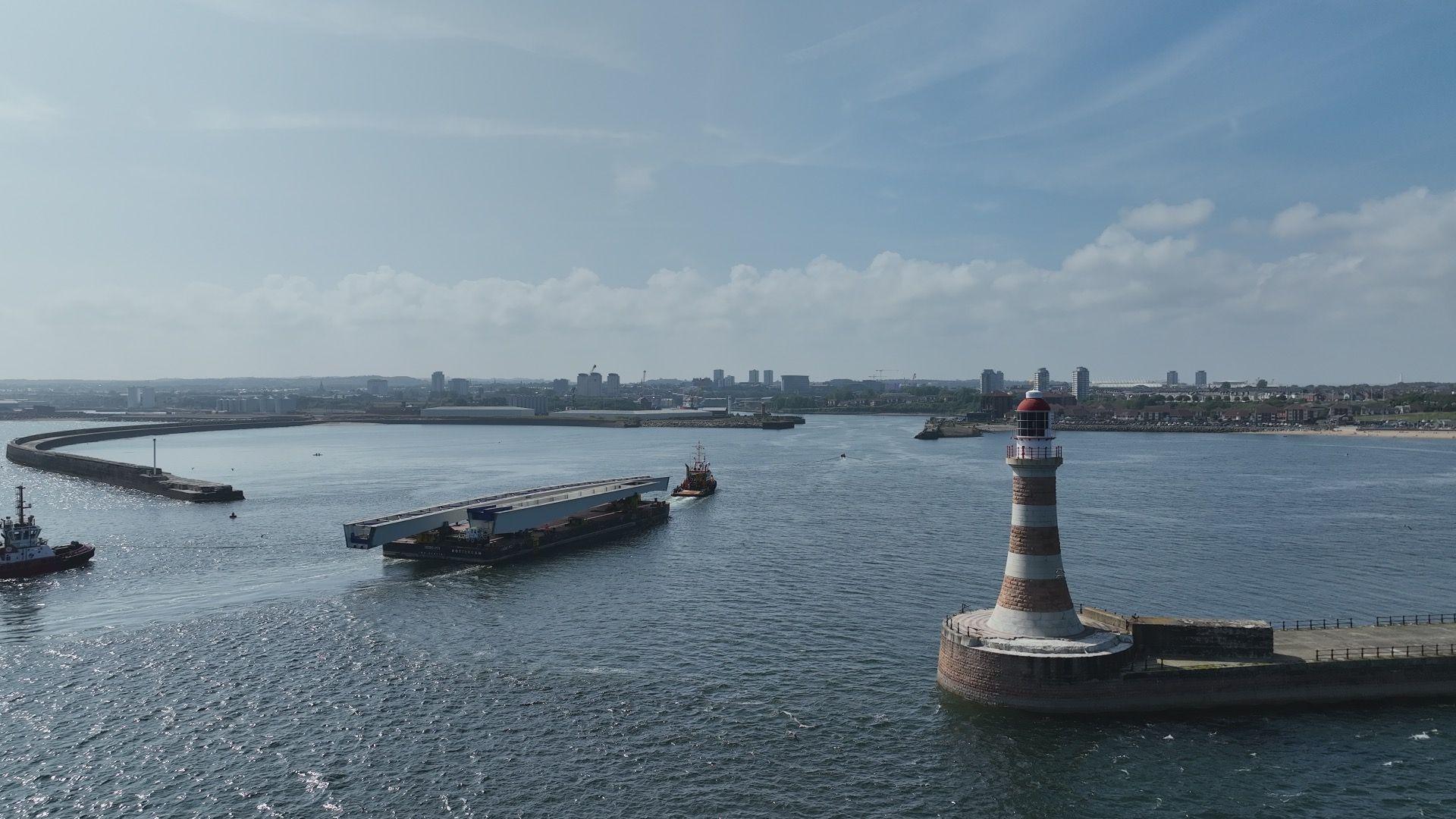 A barge carrying a metal structure through two piers. A lighthouse can be seen to the right.