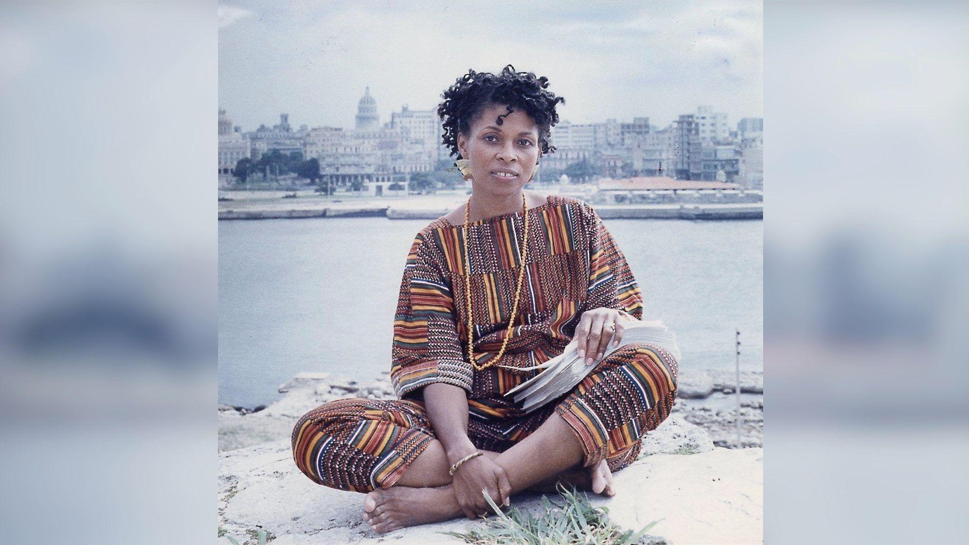 A colour image of Assata Shakur sitting on a the ground and holding the manuscript of her autobiography. The skyline of Old Havana is a body of water is behind her.