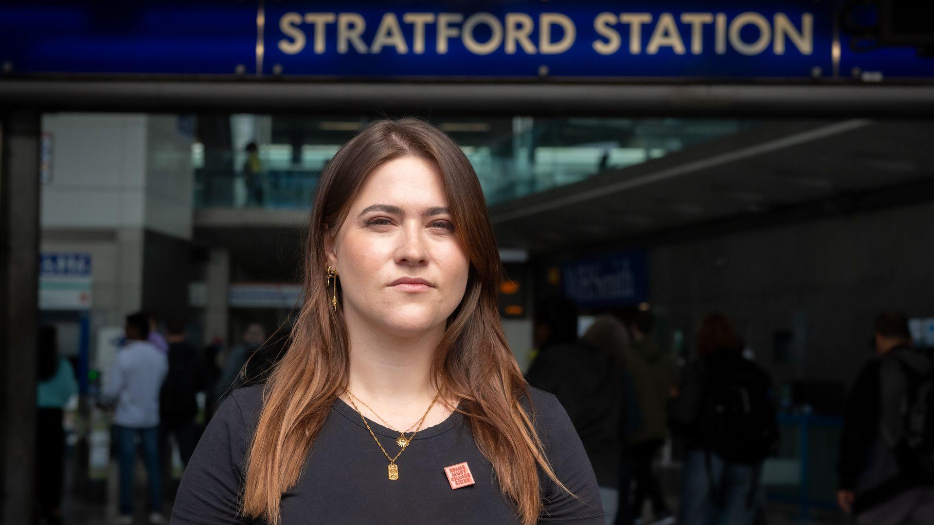 A pistillate   successful  her 30s, with agelong  brownish  hairsbreadth  successful  a achromatic  T-shirt is lasting  successful  beforehand   of the entranceway  to Stratford station. She is looking consecutive  into the camera. She is wearing a tiny  reddish  badge which says 'Shame indispensable   alteration  sides'.