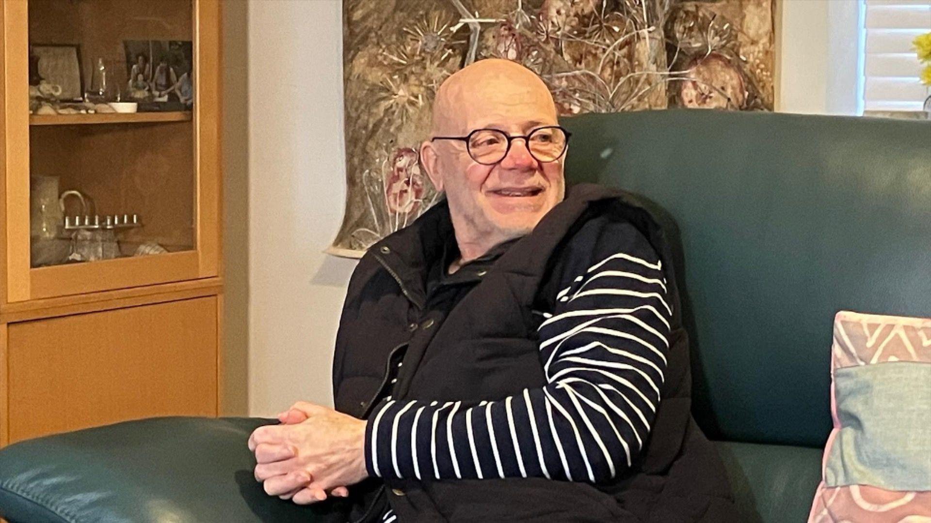 Malcolm wears a stripy navy and white top under a black gilet and sits on a green sofa. He is smiling and wears glasses.