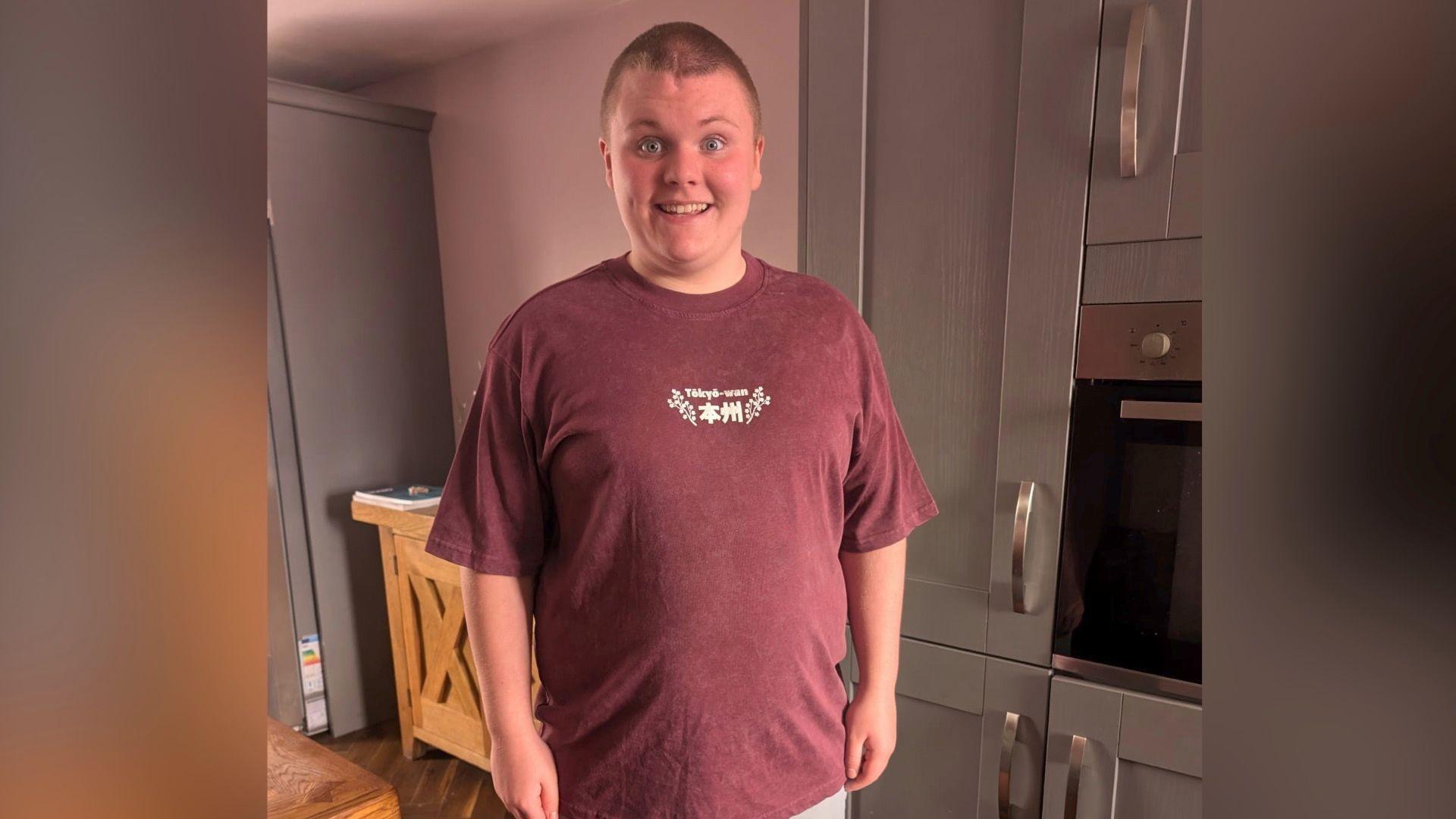 A tall teenage boy wearing purple t shirt stands and smiles at the camera. he has very short brown hair, blue eyes and in the background is a family kitchen.