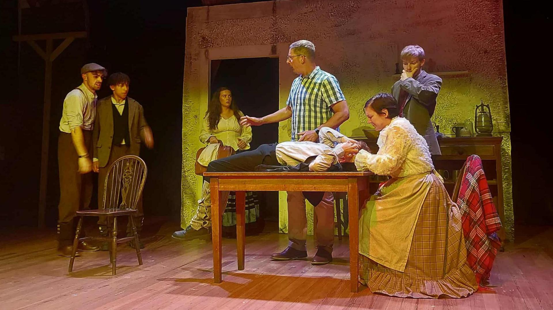 A scene from the play in which seven actors appear in a kitchen. They wear clothing from the 1830s - the men are in braces and flat caps while the women wear long skirts and aprons. One man lies on a table and holds his head, another looks on with a concerned expression.