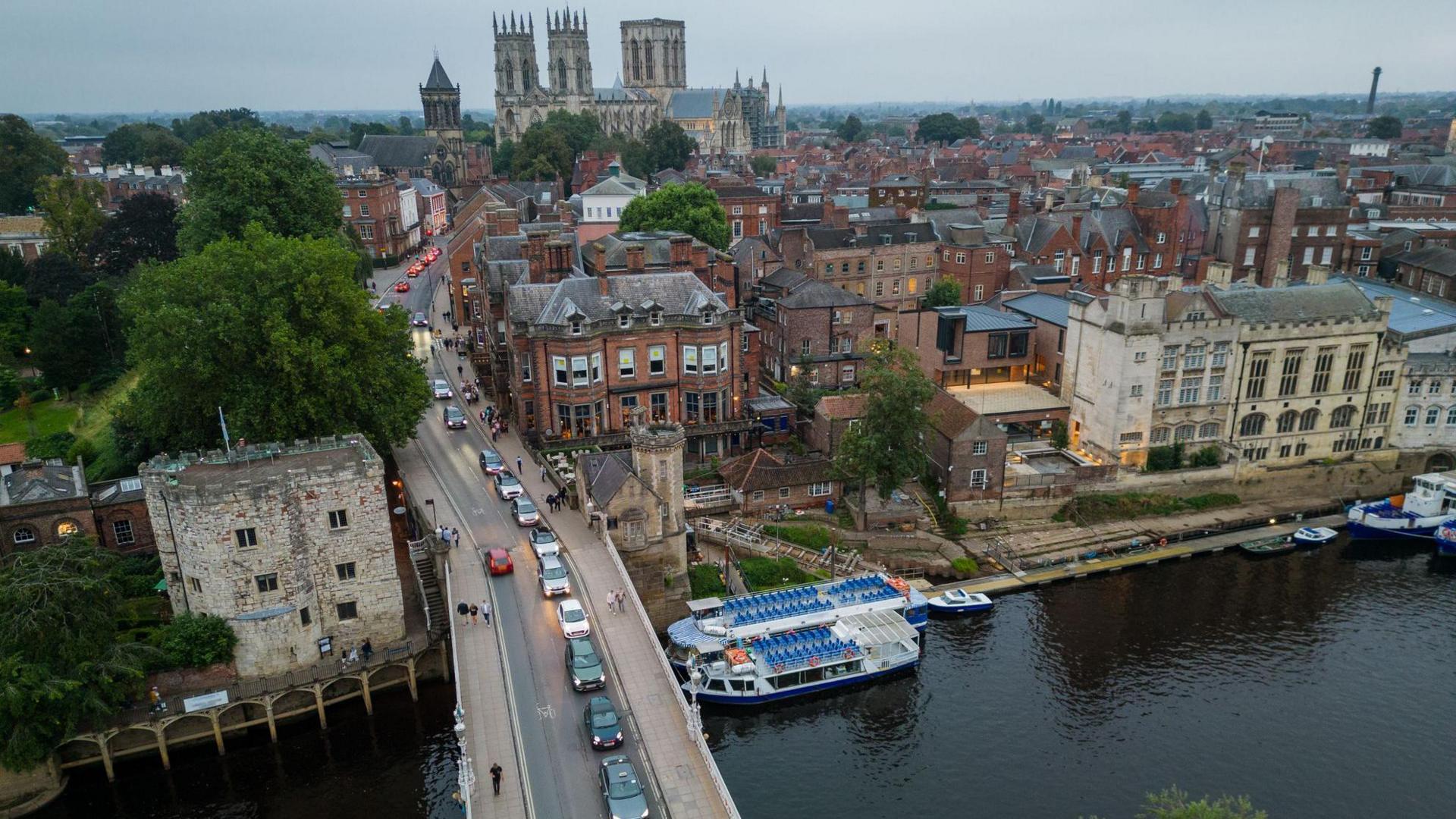 A overhead shot of Lendal Bridge in York. A bridge with a line of vehicles queued up on it. The bridge crosses a river, which has five boats moored up on the side. A view of lots of rooftops across York city centre and York Minster in the distance.