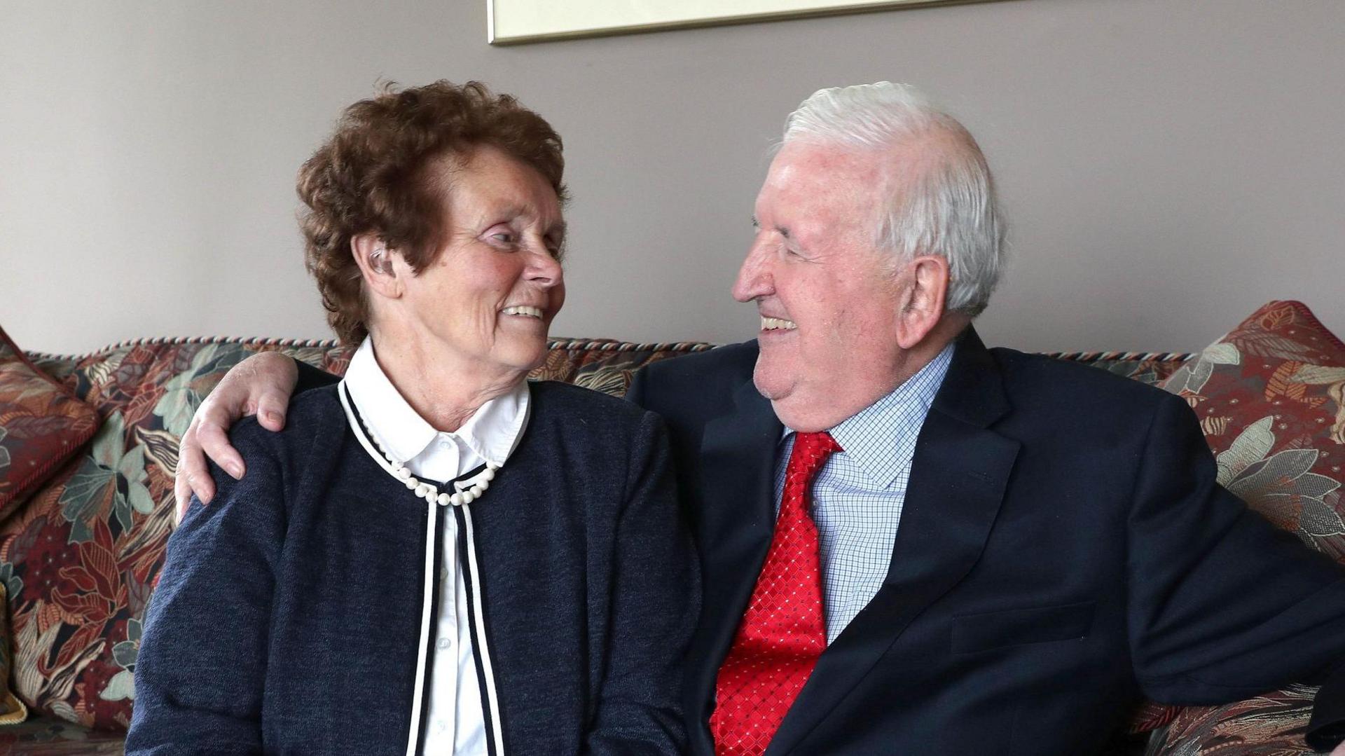 Sir Kenneth Bloomfield has grey hair and is wearing a dark suit with a red tie. He is sitting on a red and brown sofa and is smiling at his wife who has short dark hair and is wearing a white blouse with a navy cardigan and is smiling back at him. They are both in their 80s.
