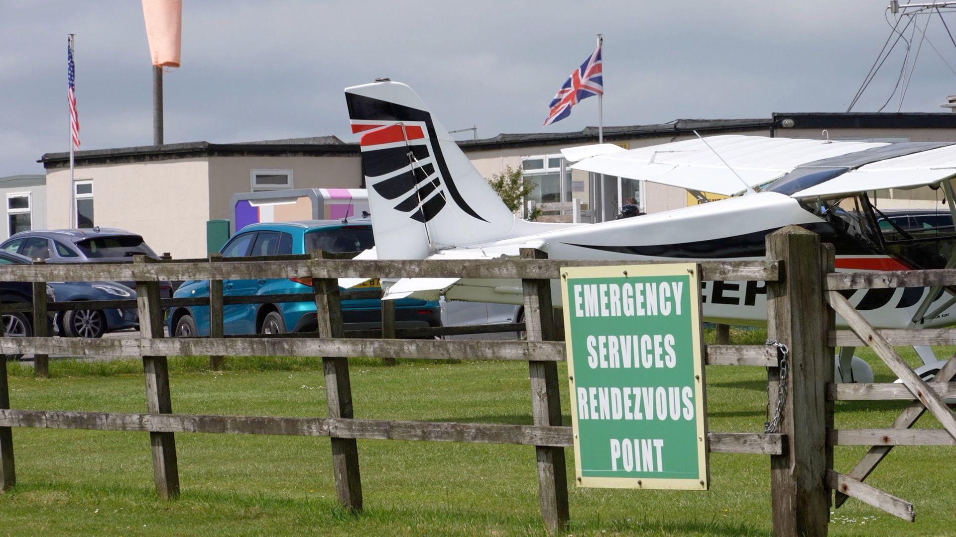 A green sign reading emergency services rendezvous point is mounted to a wooden fence. Behind the fence is a white, black and red light aircraft.