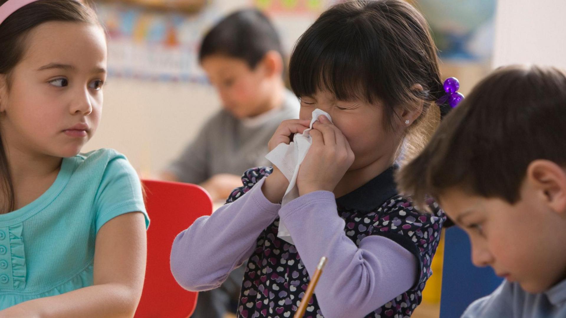A little girl sneezing into a tissue at nursery, while another child looks on