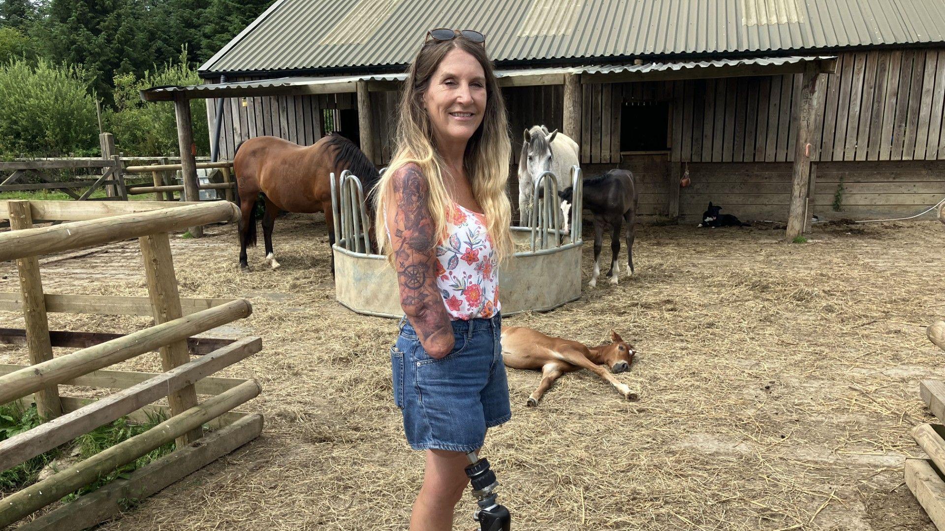 Caroline stands looking at the camera. She has long blond hair held back by sunglasses on her head. She is wearing denim shorts and a floral vest top. Caroline has a metal prosthetic leg and her arm below her elbow has been amputated. In the background are two horses and two foals, with a barn behind.