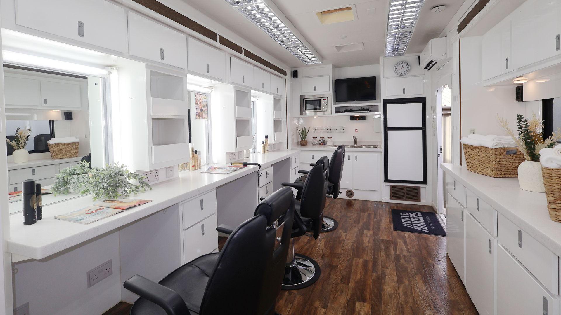 A makeup and wardrobe room inside a trailer. There are three sets of desks, each fitted with a mirror and drawers. There are makeup placed on the desks and three black swivel chairs. There is also a sink and extra drawers and cupboards on the other side of the room. 