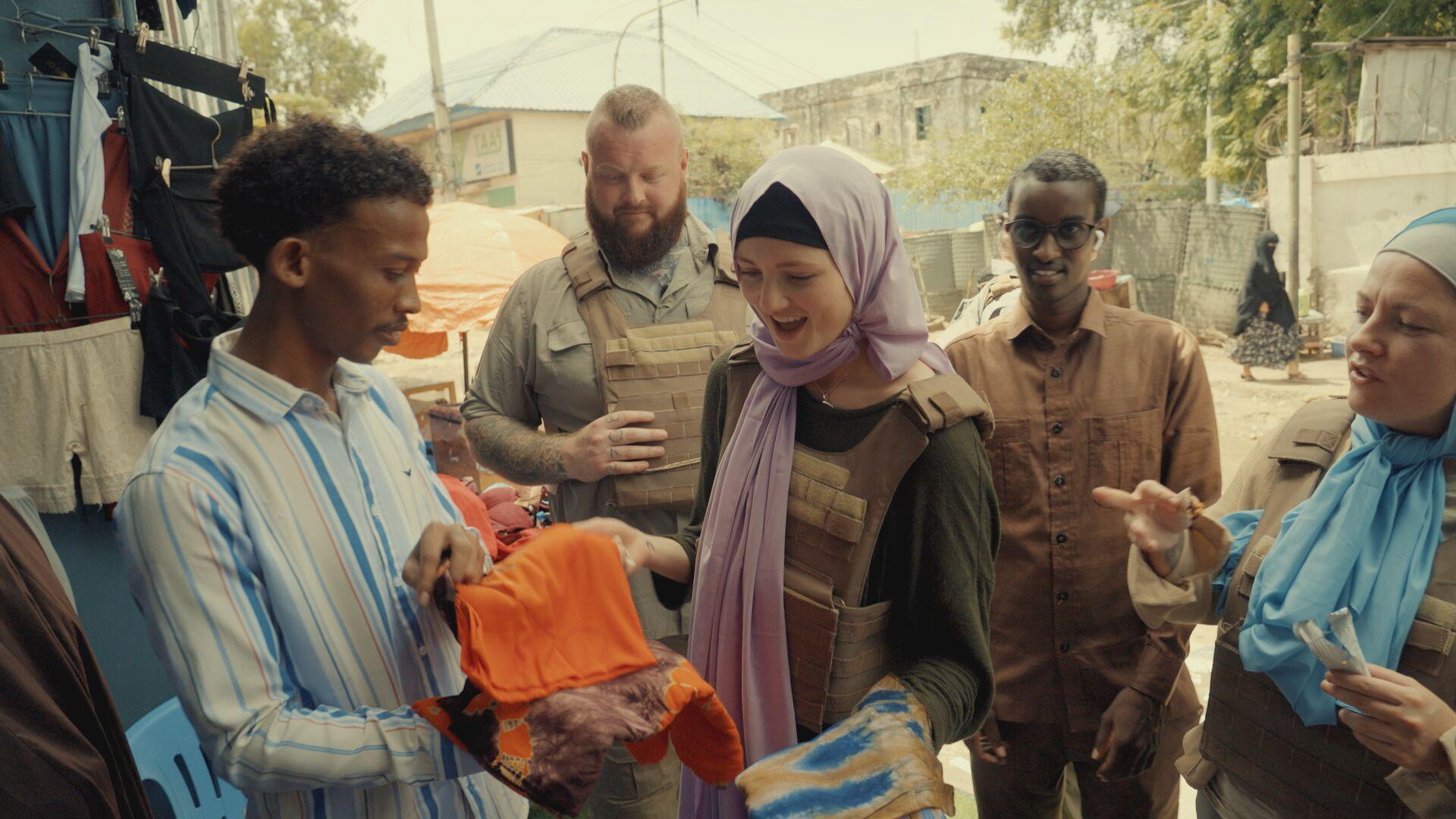 Mathilda at a market in Somalia. She's pictured wearing a stab vest and a purple head scarf while she smiles at the shop owner who holds out an orange fabric towards her. She's with fellow participants Jess and Nathan who also wear the protective vests and behind them is a sunny, sandy street. 