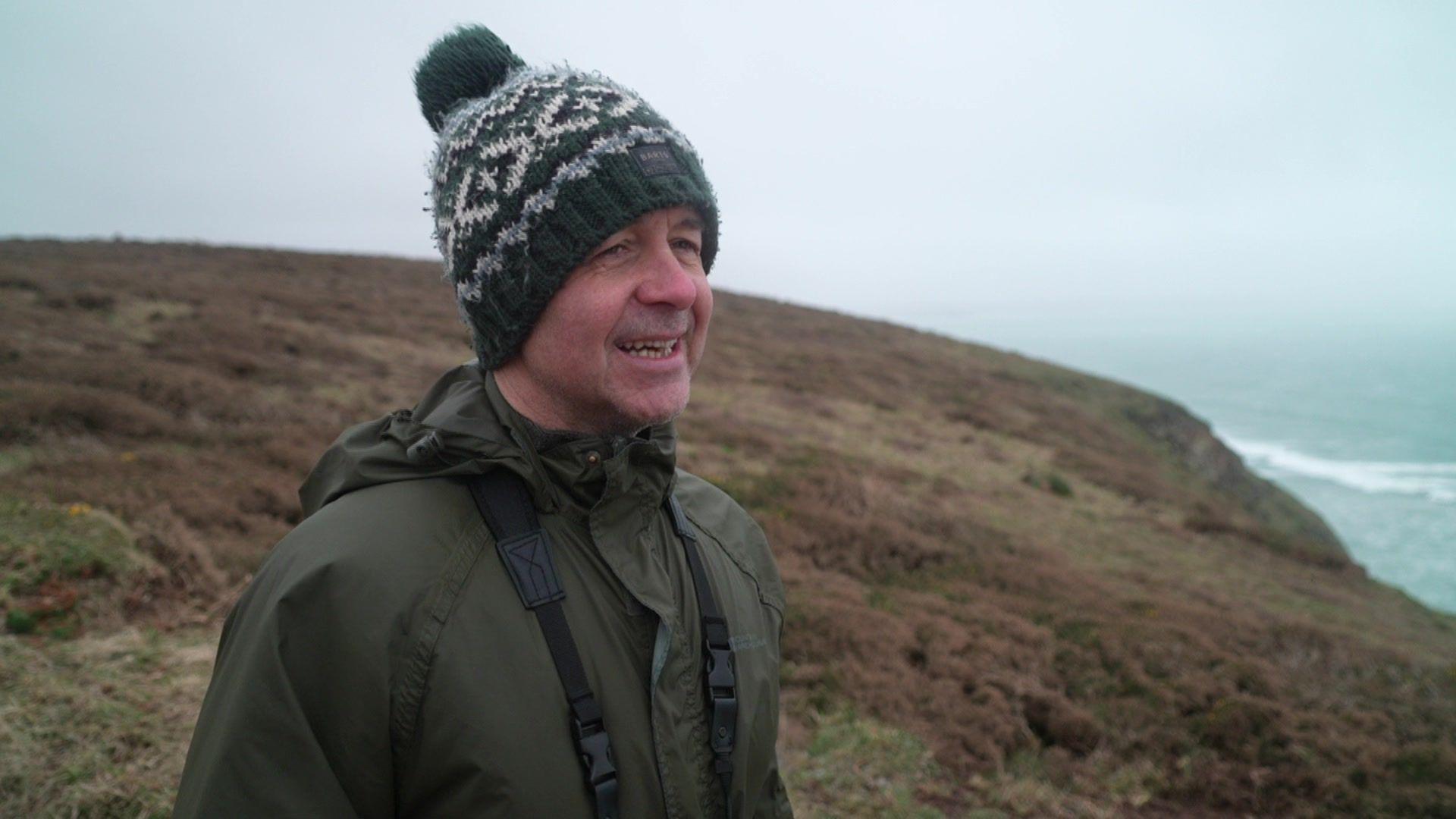 A man stood on a coast path with the sea behind him. It is a misty day and he is wearing a military green coat and a woolly hat, and also has a pair of binoculars around his neck